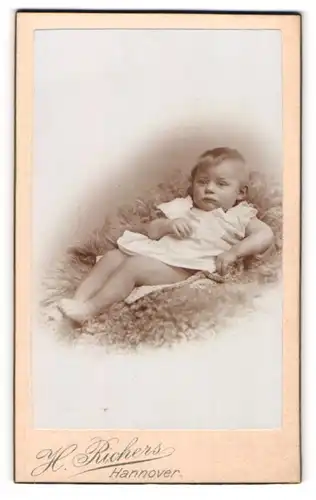 Fotografie H. Richers, Hannover, Kleinkind in weissem Kleid liegt auf einem Fell