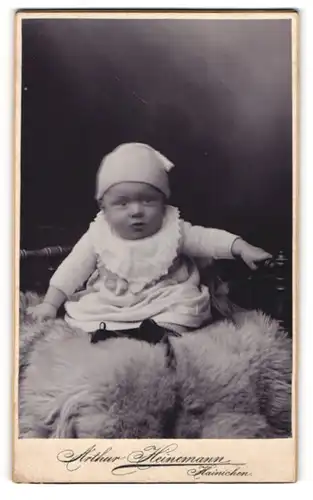 Fotografie Arthur Heinemann, Hainichen, Kleinkind mit weisser Mütze sitzt auf einem Fell