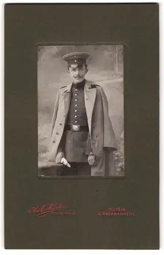 Fotografie A. & E. Hürter, Mutzig, junger Soldat in Uniform mit Mantel und Schirmmütze