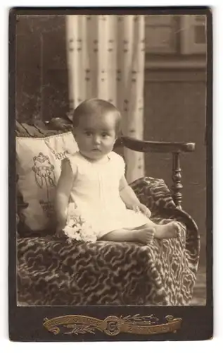 Fotografie Atelier Güttges, Zittau i / S., Portrait niedliches Kleinkind im weissen Hemd mit Blumen auf Decke sitzend