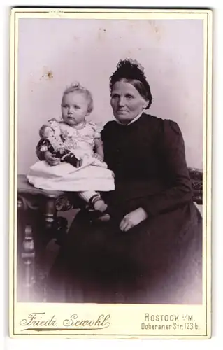 Fotografie Friedr. Servohl, Rostock i/M., Portrait Baby mit Puppe auf dem Schoss der Grossmutter sitzend
