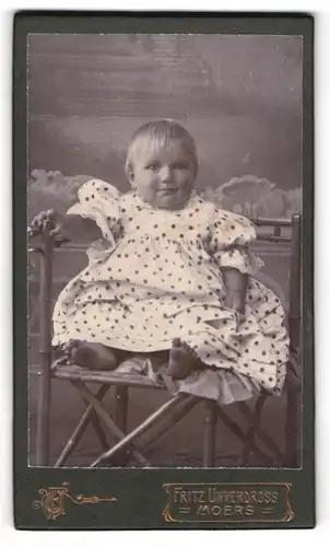 Fotografie Fritz Unverdross, Moers, Portrait süsses Kleindkind in weitem Kleidchen auf einem Stuhl posierend
