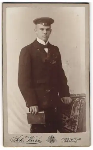 Fotografie Joh. Verra, Rosenheim, Portrait junger Herr in Anzug mit Mütze und Buch