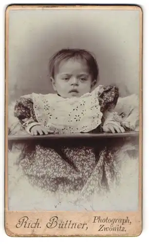 Fotografie Rich. Büttner, Zwönitz, Portrait niedliches Kleinkind im hübschen Kleid mit Latz im Hochstuhl