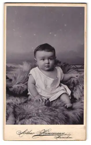 Fotografie Arthur Heinemann, Hainichen, Portrait niedliches Kleinkind im weissen Hemd auf Fell sitzend
