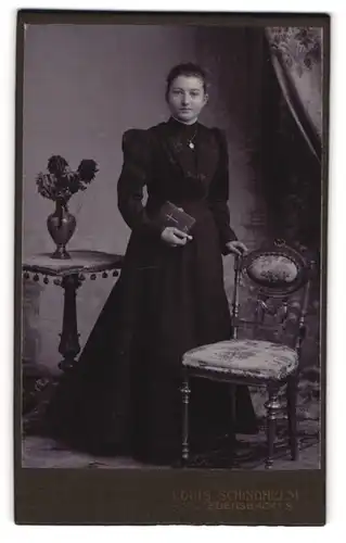 Fotografie Louis Schindhelm, Ebersbach i / S., Portrait junge Dame im schwarzen Kleid mit Buch an Stuhl gelehnt