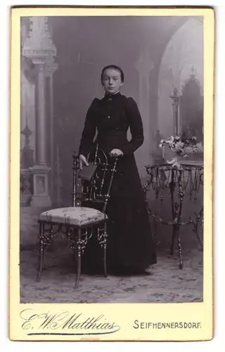 Fotografie E. W. Matthias, Seifhennersdorf, Portrait junge Dame im schwarzen Kleid mit Buch an Stuhl gelehnt