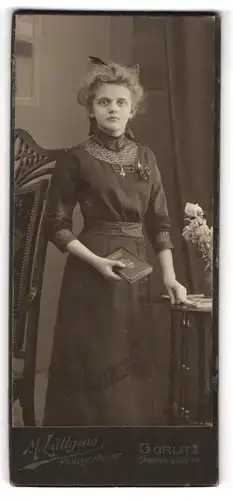 Fotografie M. Lüttgens, Görlitz, Portrait hübsch gekleidete Dame mit Buch an Tisch gelehnt