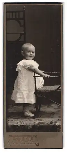 Fotografie Paul Glaue, Braunschweig, Portrait niedliches Kleinkind im weissen Kleid an Stuhl gelehnt