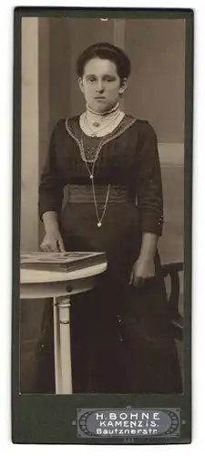 Fotografie H. Bohne, Kamenz i. S., Portrait dunkelhaarige Schönheit mit einem Fotoalbum am Tisch stehend