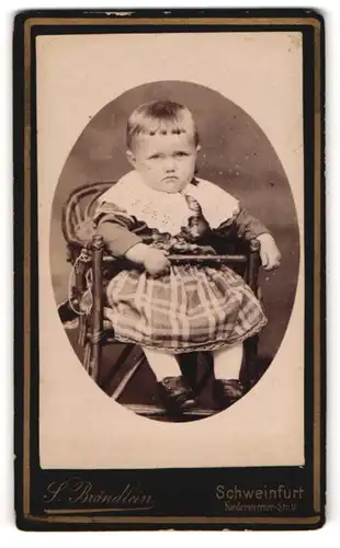 Fotografie S. Brändlein, Schweinfurt, Portrait niedliches Kleinkind im karierten Kleid im Hochstuhl sitzend