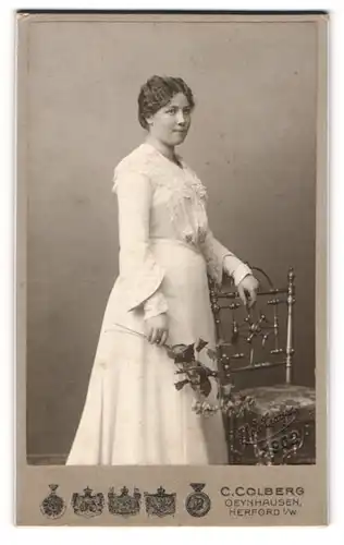 Fotografie C. Colberg, Oeynhausen, Portrait junge Dame im weissen Kleid mit Blume an Stuhl gelehnt