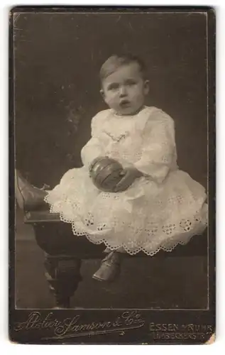 Fotografie Samson & Co., Essen a / Ruhr, Portrait niedliches Kleinkind im weissen Kleid mit Ball auf Tisch sitzend