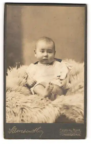 Fotografie Samson & Co., Essen a / d Ruhr, Portrait niedliches Baby im weissen Hemd auf Fell sitzend