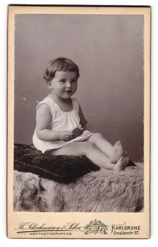 Fotografie Th. Schuhman & Sohn, Karlsruhe, Portrait niedliches Kleinkind im weissen Hemd mit Keks auf Kissen sitzend