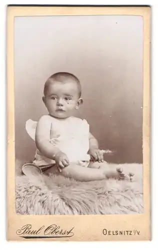 Fotografie Paul Oberst, Oelsnitz i. V., Portrait niedliches Baby im weissen Hemd auf Fell sitzend