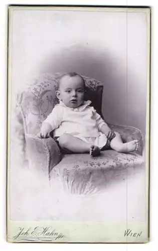 Fotografie Joh. E. Hahn, Wien, Portrait niedliches Baby in weissen Hemdchen im Sessel lehnend