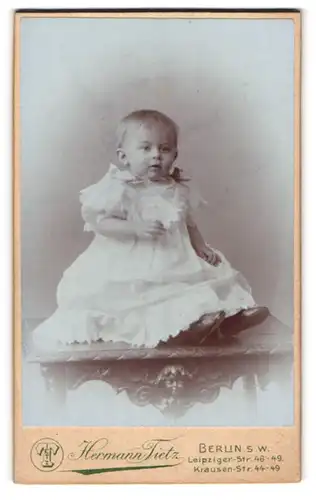 Fotografie Hermann Tietz, Berlin-SW, Portrait niedliches Kleinkind im weissen Kleid auf Tisch sitzend
