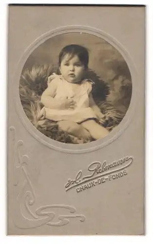 Fotografie H. Rebmann, Chaux-de-Fonds, Portrait niedliches Baby im weissen Hemd auf Fell sitzend