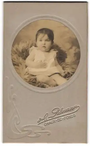 Fotografie H. Rebmann, Chaux-de-Fonds, Portrait niedliches Baby im weissen Hemd auf Fell sitzend