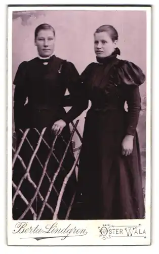 Fotografie Berta Lindgren, Öster Wala, Portrait zwei junge Damen in schwarzen Kleidern an Zaun gelehnt