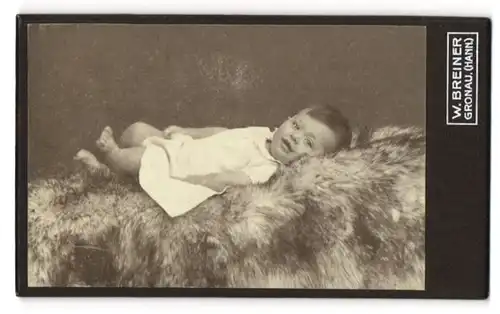 Fotografie W. Breiner, Gronau i. Hann., Portrait lachendes Baby im weissen Kleidchen auf Fell liegend