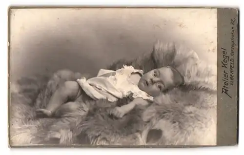 Fotografie Atelier Kegel, Elberfeld, süsses Baby im weissen Kleidchen auf Fell liegend