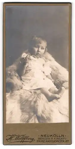 Fotografie H. Wolffberg, Berlin-Neukölln, Portrait niedliches Kleinkind im weissen Kleid auf Fell sitzend