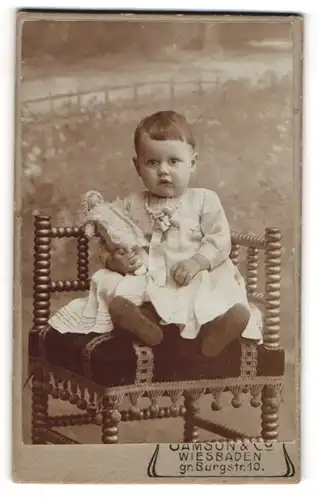 Fotografie Samson & Co., Wiesbaden, Portrait niedliches Kleinkind im hübschen Kleid mit Puppe auf Stuhl sitzend