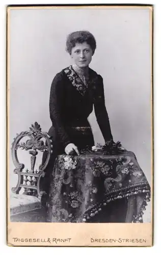 Fotografie Taggeselle & Ranft, Dresden, junger Frau Grete im dunklen Kleid am Tisch stehend, 1901