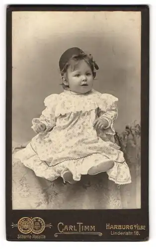 Fotografie Carl Timm, Harburg a / Elbe, Portrait süsses Kleinkind im hübschen Kleid auf Tisch sitzend
