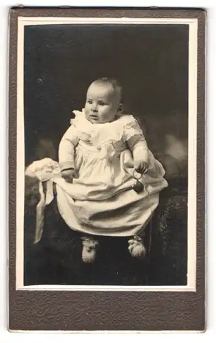 Fotografie unbekannter Fotograf und Ort, Portrait niedliches Baby im weissen Kleid mit Rassel auf Fell sitzend