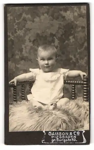 Fotografie Samson & Co., Wiesbaden, Portrait niedlich blickendes Kleinkind auf Fell sitzend