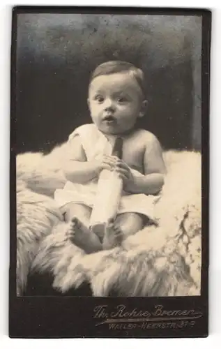 Fotografie Th. Rohse, Bremen, Portrait niedliches Baby im weissen Hemd mit Trinkflasche auf Fell sitzend