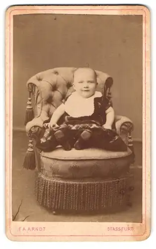 Fotografie F. A. Arndt, Stassfurt, Portrait niedliches Kleinkind im modischen Kleid mit Latz auf Sessel sitzend