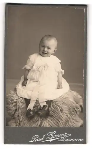 Fotografie Paul Lorenz, Wolmirstedt, Portrait niedliches Kleinkind im weissen Kleid auf Fell sitzend