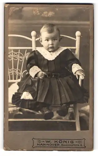 Fotografie W. König, Hannover, Portrait niedliches Kleinkind im Spitzenkleid auf Kissen sitzend