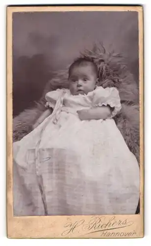 Fotografie H. Richers, Hannover, Portrait niedliches Baby im weissen Kleid auf Fell sitzend