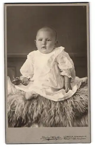 Fotografie Atelier Woelfer, Lübeck, Portrait niedliches Kleinkind im weissen Taufkleidchen auf Fell sitzend