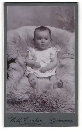 Fotografie Wilh. Everhan, Gummersbach, Portrait niedliches Baby im weissen Hemd auf Fell sitzend