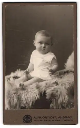Fotografie Alfr. Ortloff, Ansbach, Portrait niedliches Kleinkind im weissen Hemd auf Fell sitzend