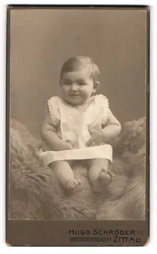 Fotografie Hugo Schröder, Zittau, Portrait niedliches Kleinkind im weissen Hemd auf Fell sitzend