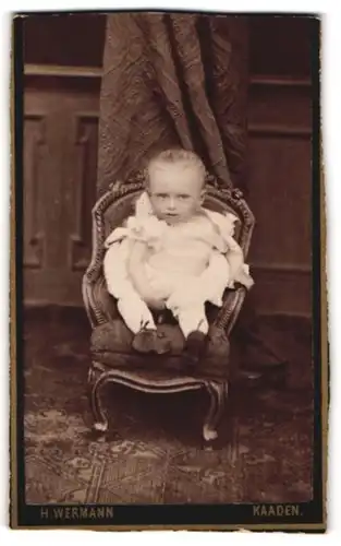 Fotografie H. Wermann, Kaaden, Baby im weissen Kleid sitzend auf einem Stuhl