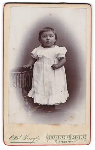 Fotografie Otto Graef, Oldenburg, Portrait niedliches Kleinkind im weissen Kleid auf Stuhl stehend