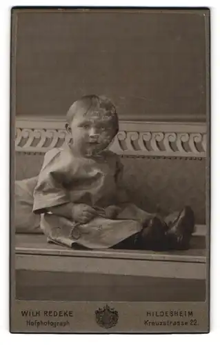 Fotografie Wilh. Redeke, Hildesheim, Portrait niedliches Kleinkind im hübschen Kleid auf Bank sitzend
