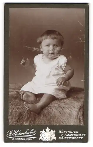 Fotografie J. Heimhuber, Immenstadt, Portrait niedliches Kleinkind im weissen Hemd auf Fell sitzend