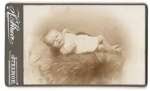 Fotografie H. Struve, Itzehoe, Portrait niedliches Baby im weissen Hemd auf Fell liegend