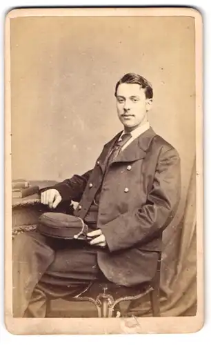 Fotografie Henry Death, London-SE, Portrait bürgerlicher Herr mit Schirmmütze am Tisch sitzend