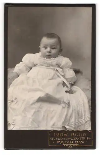 Fotografie Ludw. Kühn, Berlin-Pankow, Portrait niedliches Baby im weissen Kleid auf Fell sitzend