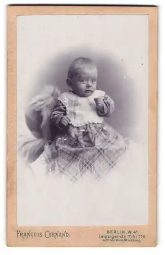 Fotografie Francois Cornand, Berlin-W, Portrait niedliches Baby im karierten Kleid auf Fell sitzend